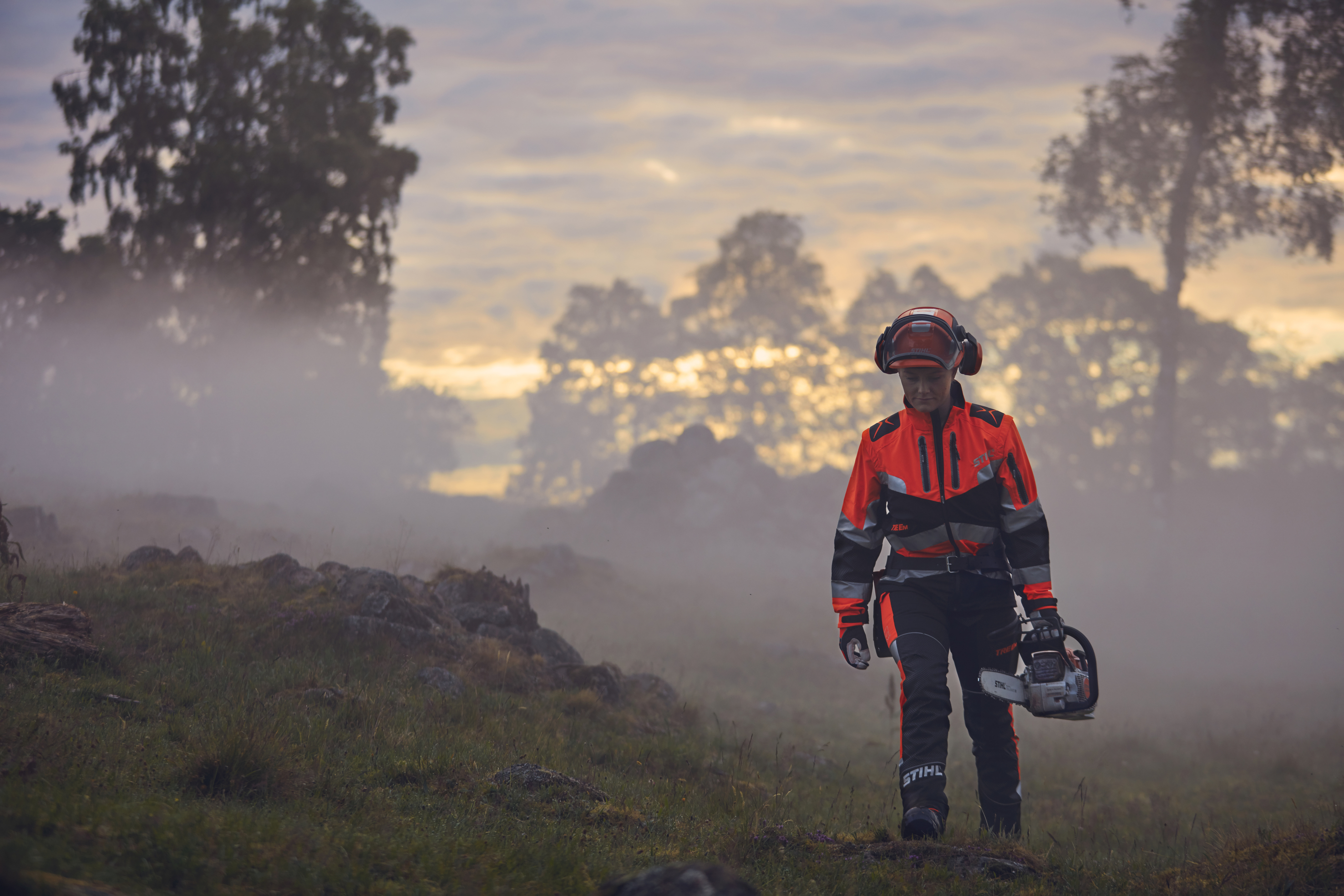 Naispuolinen metsätyöntekijä kävelee STIHL-työvaatteissa auringonnousussa mäellä