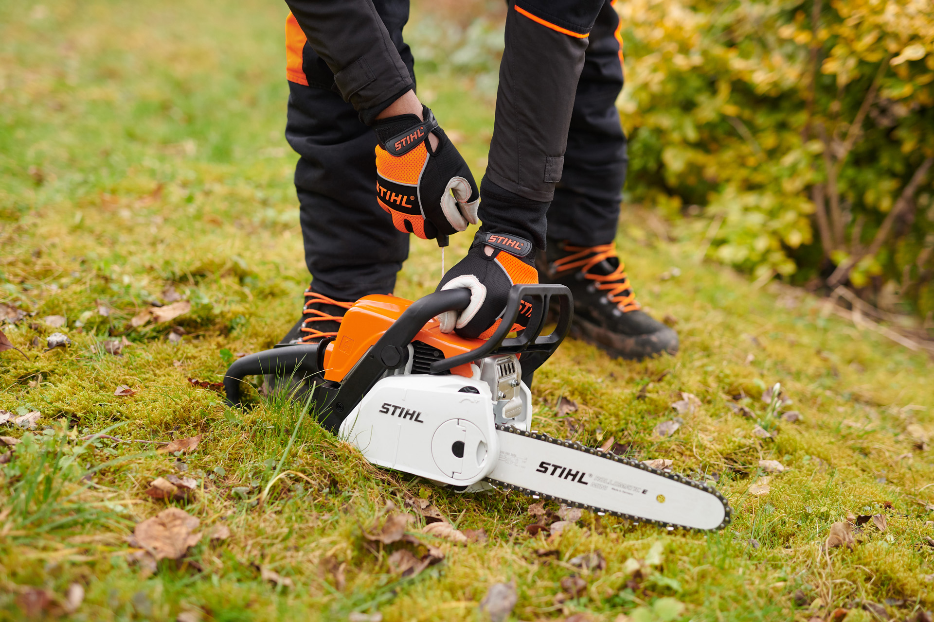 STIHL MS 180 C-BE -moottorisaha maassa ja suojakäsineitä käyttävä henkilö pitelemässä kahvasta.