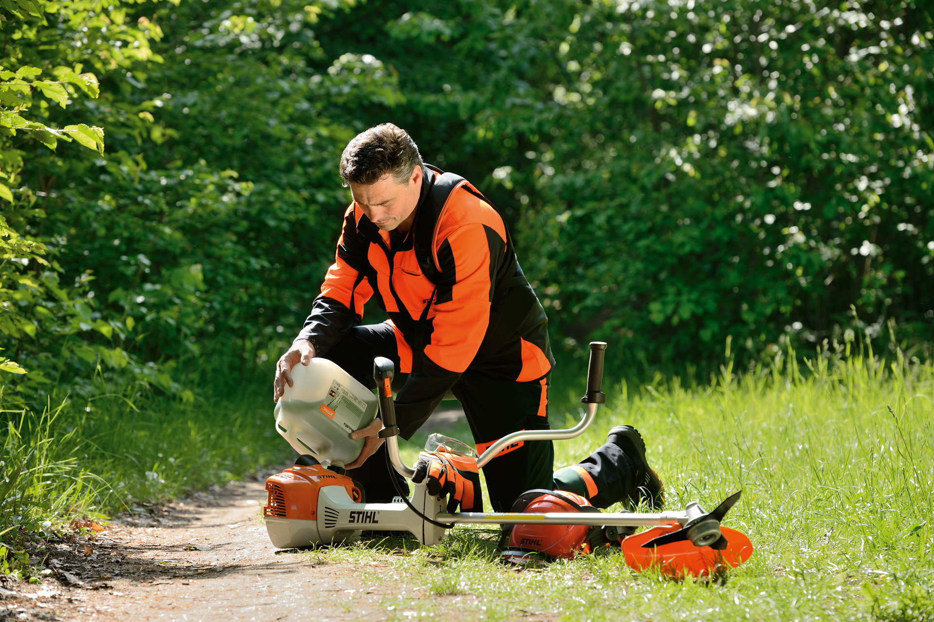 Mies täyttämässä STIHL-ruohotrimmerin polttoainesäiliötä suuresta pullosta, taustalla pensaita