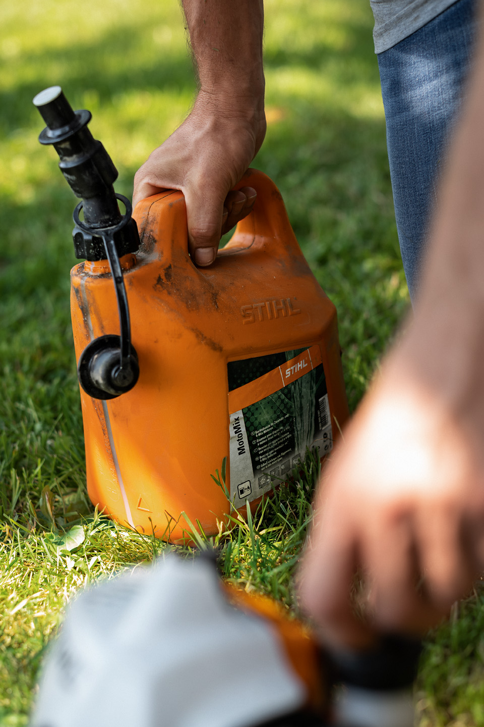 Likainen STIHL MotoMix -polttoaineseoksen kanisteri ruoholla, etualalla työkalun polttoainekorkkia avaava käsi