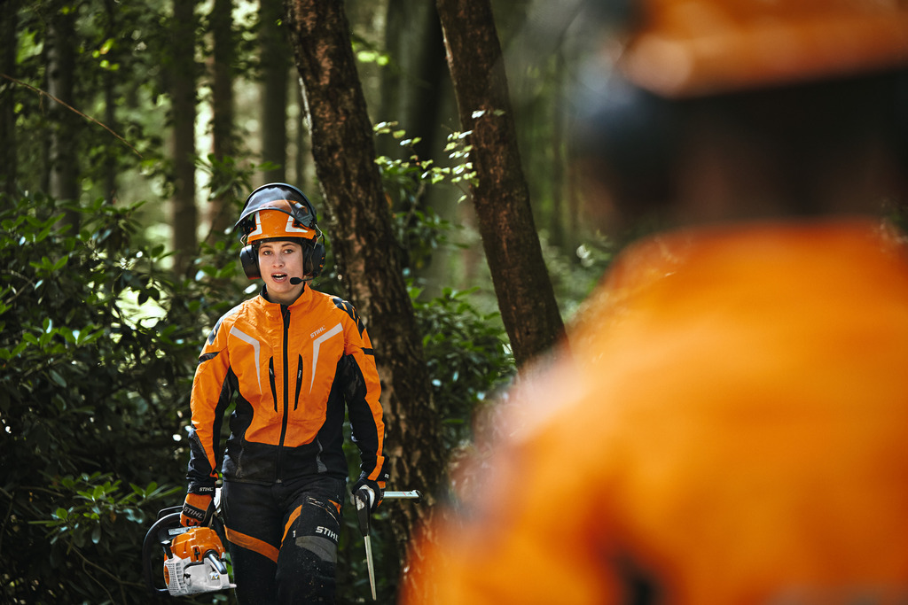 Naispuolinen metsätyöntekijä kävelee STIHL-työvaatteissa auringonnousussa mäellä