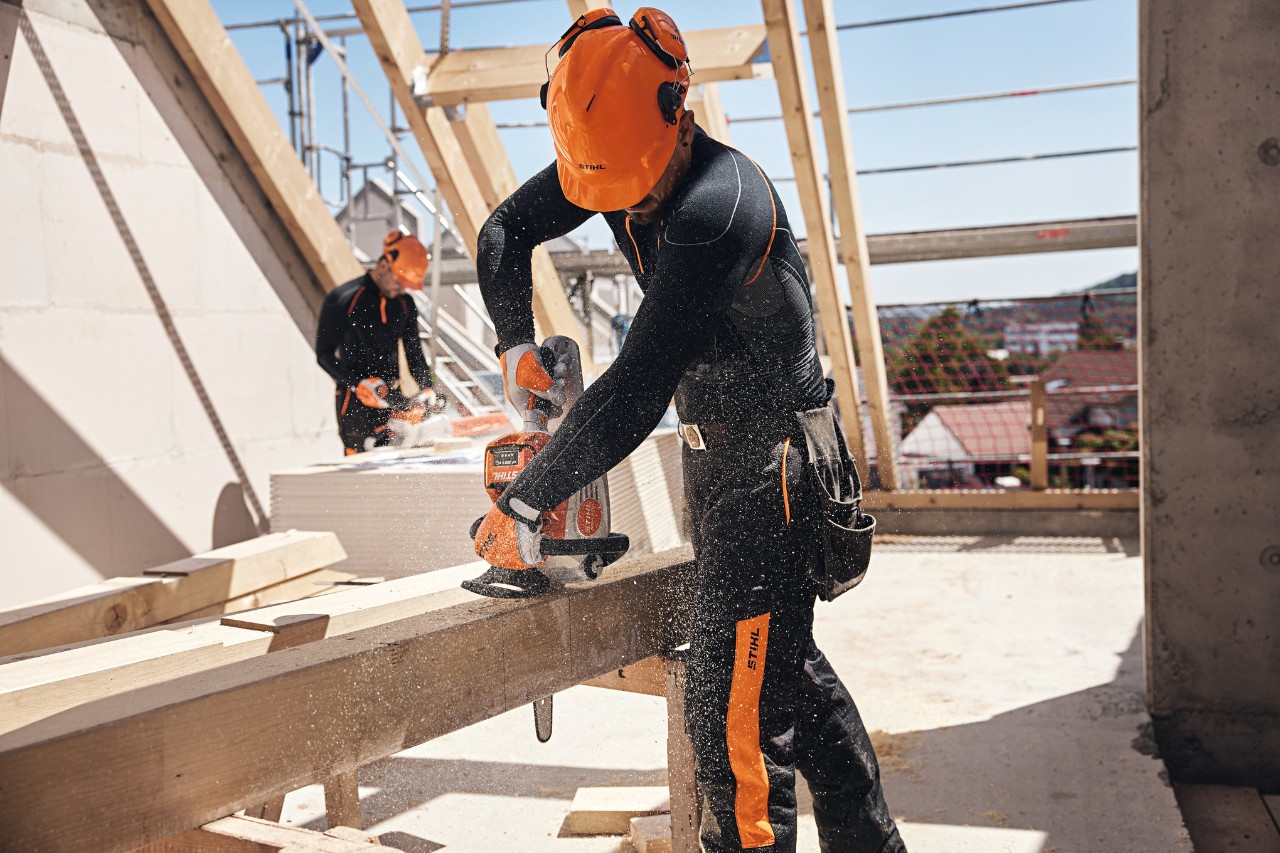 STIHL-ammattilainen sahaamassa ullakolla puupalkkia akkumoottorisahalla, toinen työntekijä käyttämässä taustalla akkumoottorisahaa