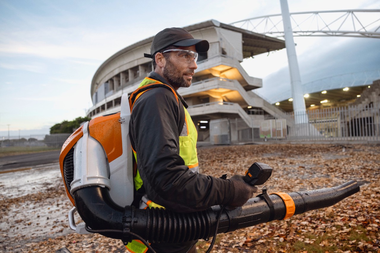 Myöhään illalla henkilö siivoaa lehtiä ja roskia stadionilta ALLPRO STIHL BRA 600 -akkulehtipuhaltimella.