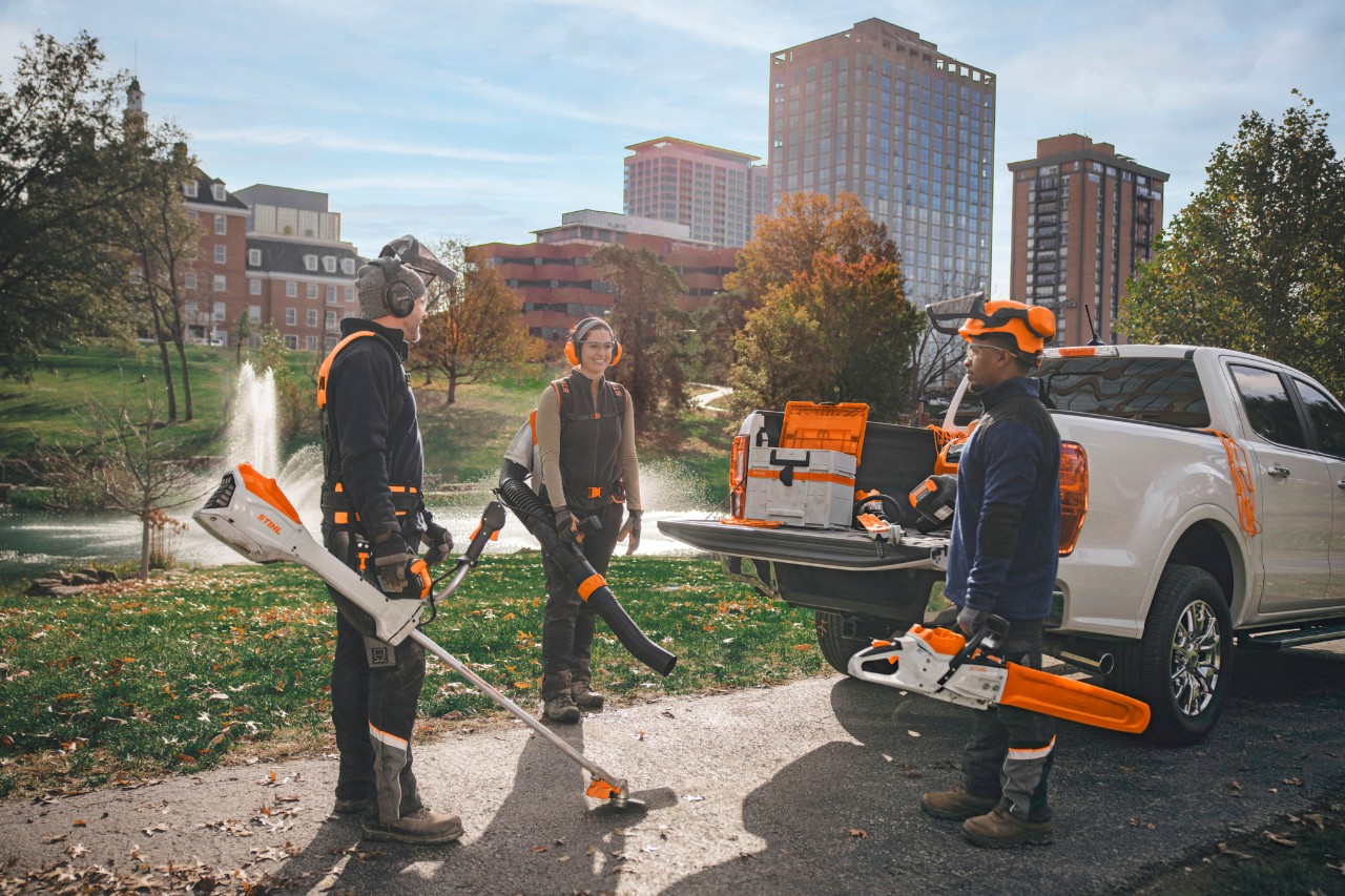 Ryhmä ammattimaisia ​​maisemanhoitajia seisoo ajoneuvojensa ympärillä ja valmistautuu työhön STIHL-akkutyökaluilla.