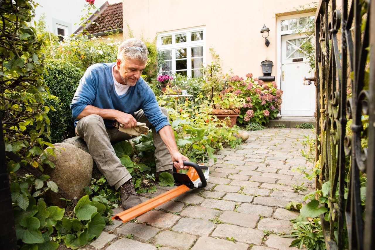 Mies istumassa suurella kivellä sähkötoimisen STIHL AI Series HSA 45 -pensasleikkurin kanssa talon edessä