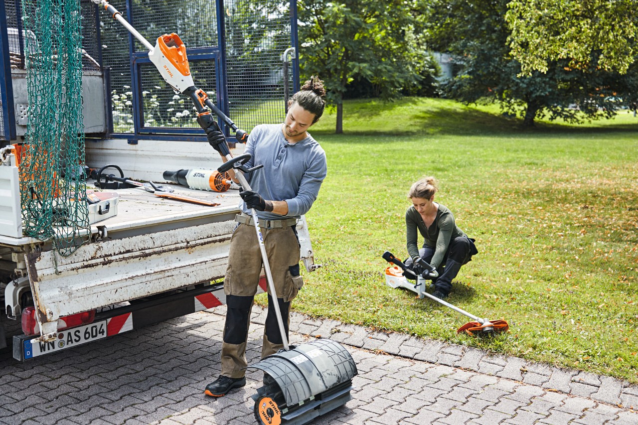 Ein Mann und eine Frau hantieren mit STIHL Akku-Geräten neben einem Fahrzeug mit offener Ladefläche, im Hintergrund eine Wiese