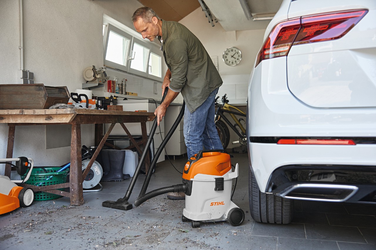 Ein Mann saugt in einer Garage mit einem STIHL Nass- und Trockensauger den Boden, neben ihm eine Werkbank und ein weißes Auto